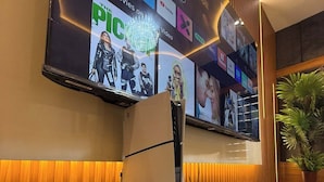 Un moderno vestíbulo de hotel con un televisor de pantalla plana grande que muestra varios servicios de streaming, un sillón de masajes y un mostrador de recepción de madera.
