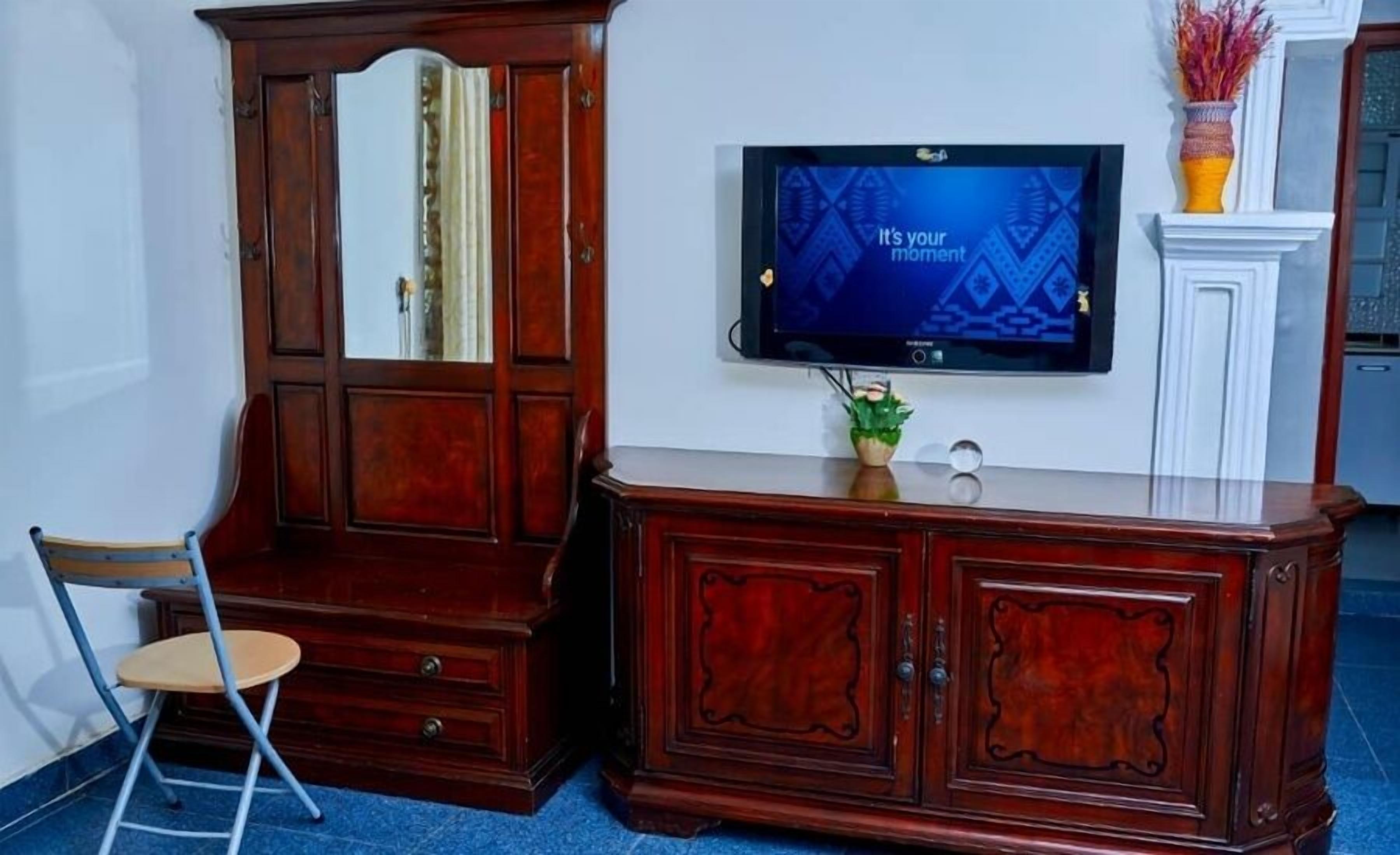 A room with a wooden cabinet, a television mounted on the wall, a small plant on the cabinet, and a chair.