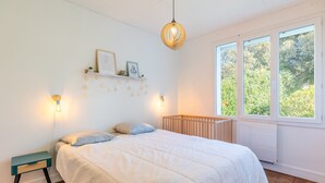 A bedroom with a bed, bedside table, wall-mounted shelf, and a window with a view of greenery. - Family home in central Fromentine with terrace, Wi-Fi, air conditioning, €800 security deposit (La Barre-de-Monts)