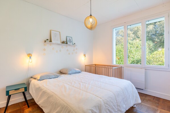 A bedroom with a bed, bedside table, wall-mounted shelf, and a window with a view of greenery.