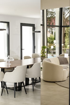 A modern dining area with a wooden table and chairs, large windows, and a view of greenery outside.
