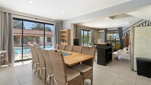 Een ruime eetruimte met een grote tafel en rieten stoelen, een keuken met een stenen muur, en uitzicht op een zwembad.