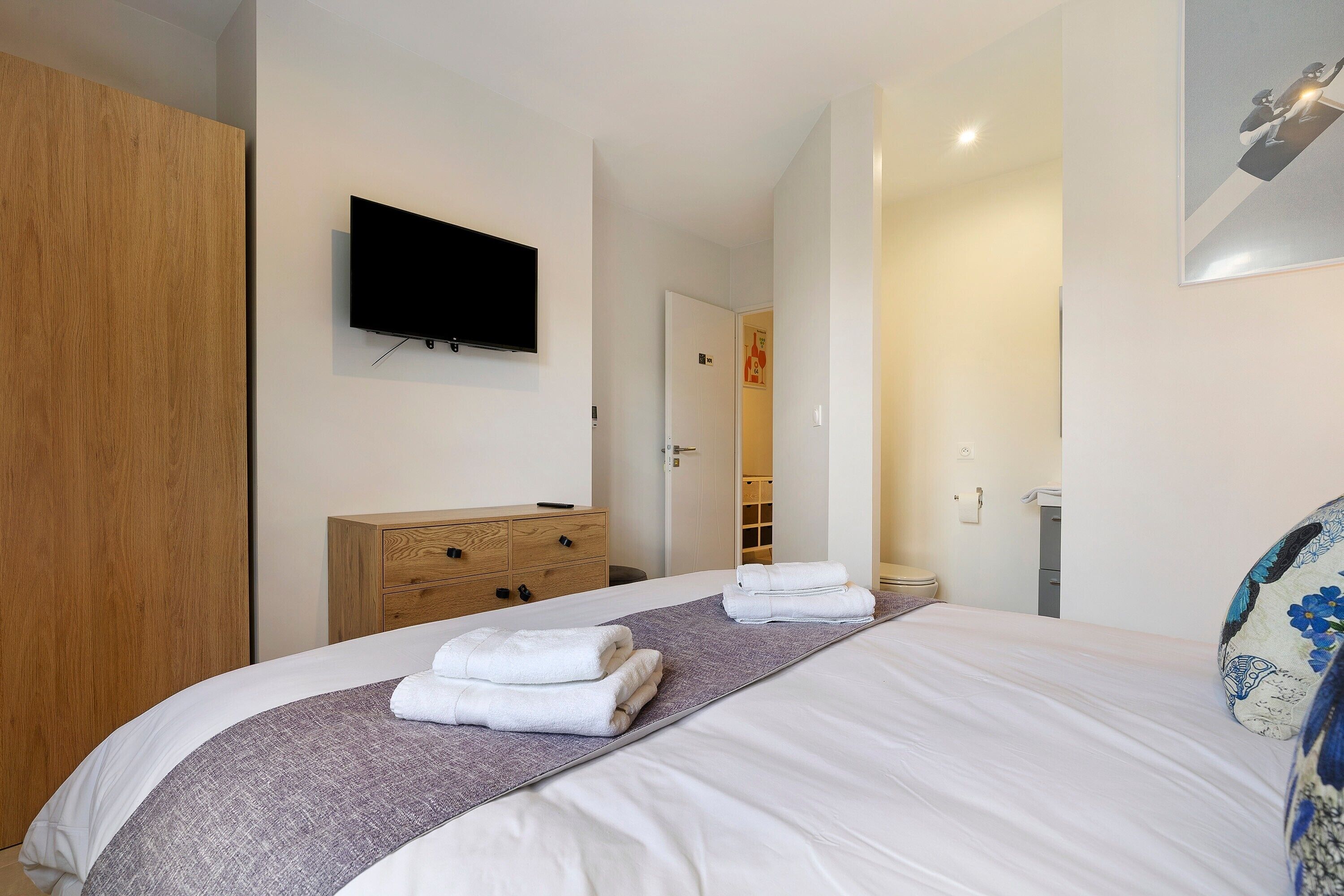 Ein Hotelzimmer mit einem Bett, einem Flachbildfernseher, einem Holzschrank, einem Badezimmer mit Toilette und Waschbecken sowie einer Tür zu einem weiteren Raum.