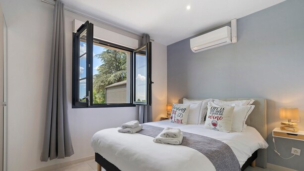 Een slaapkamer met een bed, een raam met gordijnen, een nachtkastje met een lamp en een aan de muur bevestigde airconditioning.
