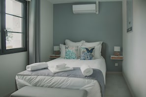 Ein Schlafzimmer mit einem Bett, Kissen, einem Fenster, einer Klimaanlage, Nachttischen und einem Beistelltisch.