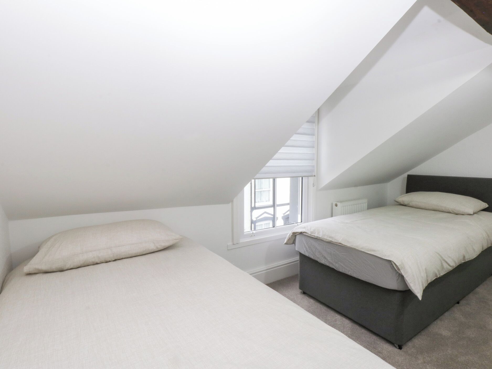 Une petite chambre mansardée, bien éclairée, avec un plafond en pente, une fenêtre et un lit doté d’une tête de lit sombre.