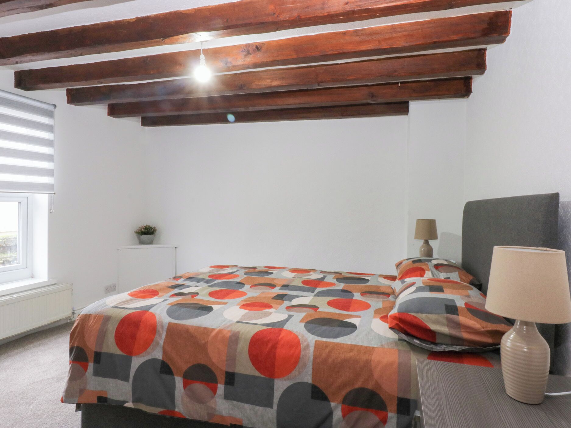 Un dormitorio con cama, mesita de noche con lámpara, ventana con persianas y viga de madera en el techo.