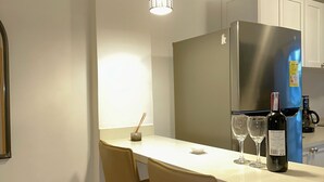 Une cuisine moderne avec un plan de travail blanc, deux tabourets de bar, un réfrigérateur en acier inoxydable et des luminaires suspendus.