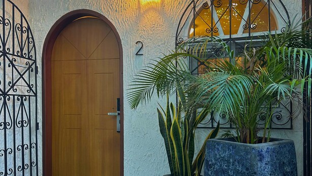 Une entrée d’immeuble avec une porte en bois, une plaque portant le numéro « 2 », un portail en fer forgé et deux grands pots de plantes.