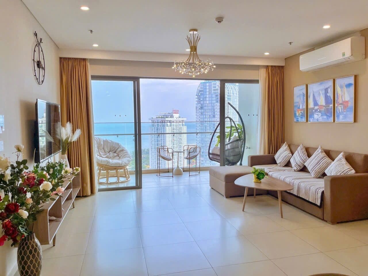 A spacious living room with a sofa, coffee table, and a TV. There are large windows with a view of buildings and a balcony with chairs.