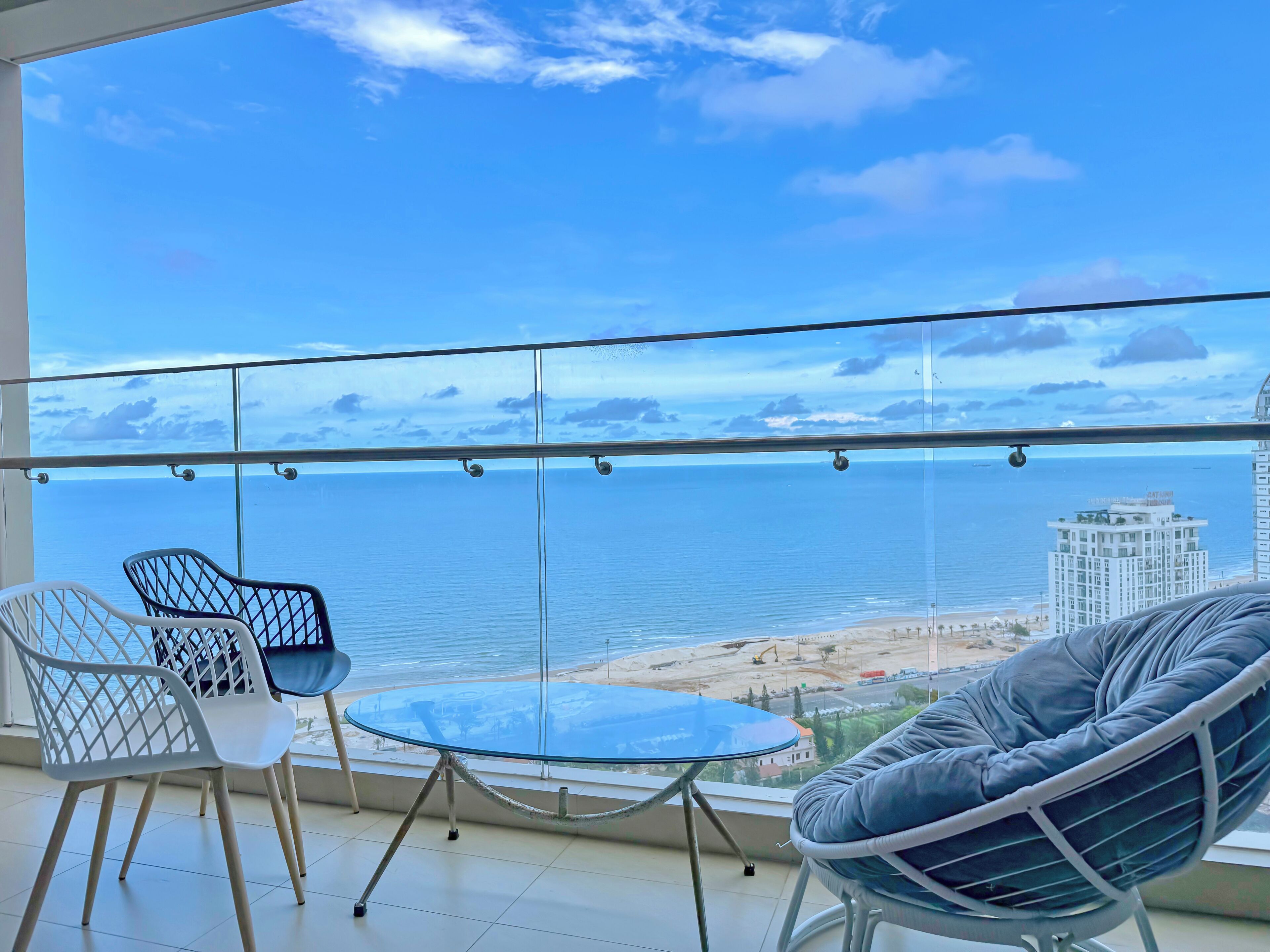 Un balcon avec une table et des chaises en verre, donnant sur une plage et l’océan.