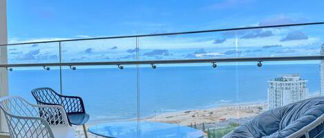 Un balcon avec une table et des chaises en verre, donnant sur une plage et l’océan.