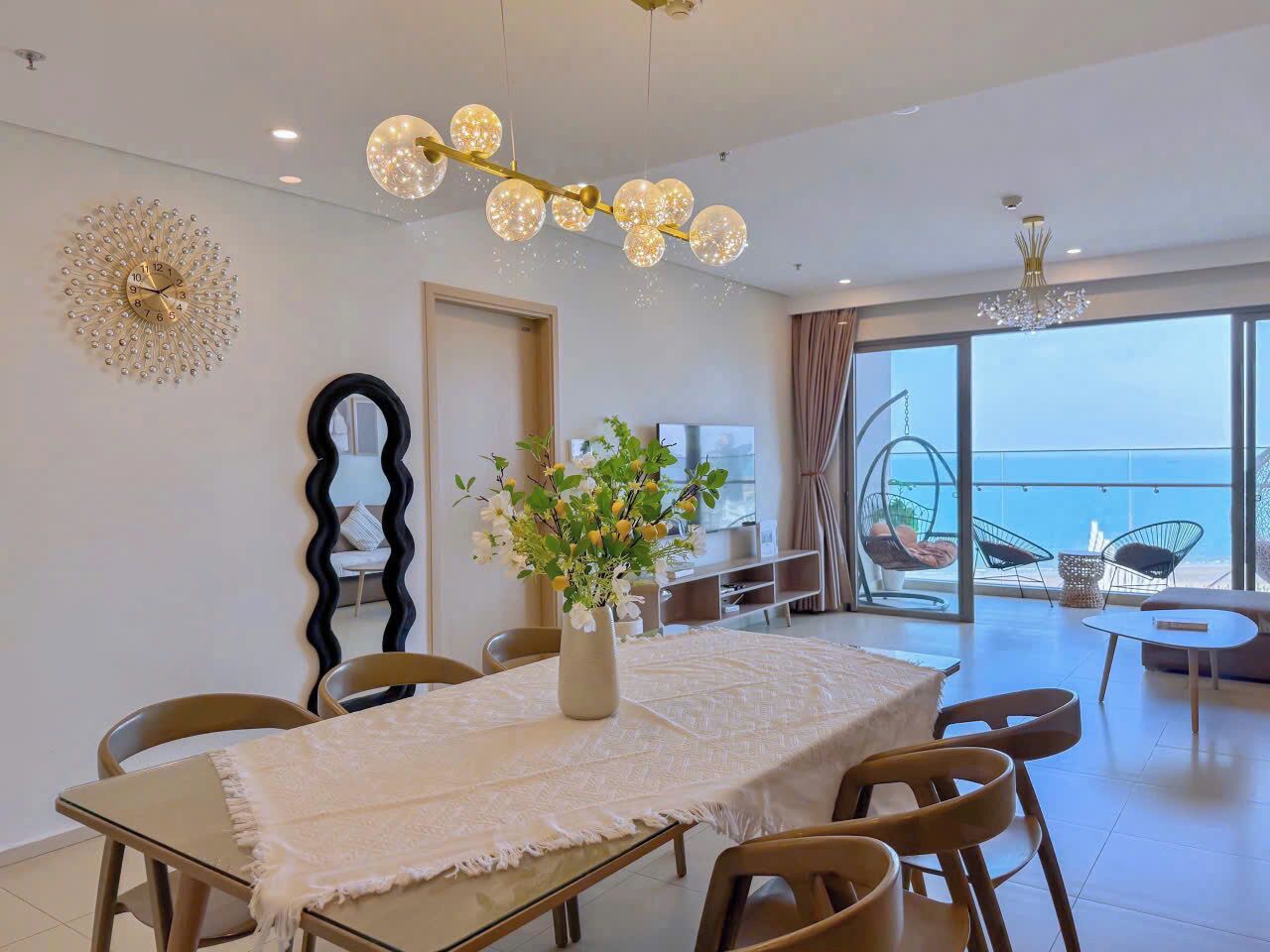 A dining area with a table, chairs, a vase with flowers, a decorative wall piece, and a view of the sea through large windows.
