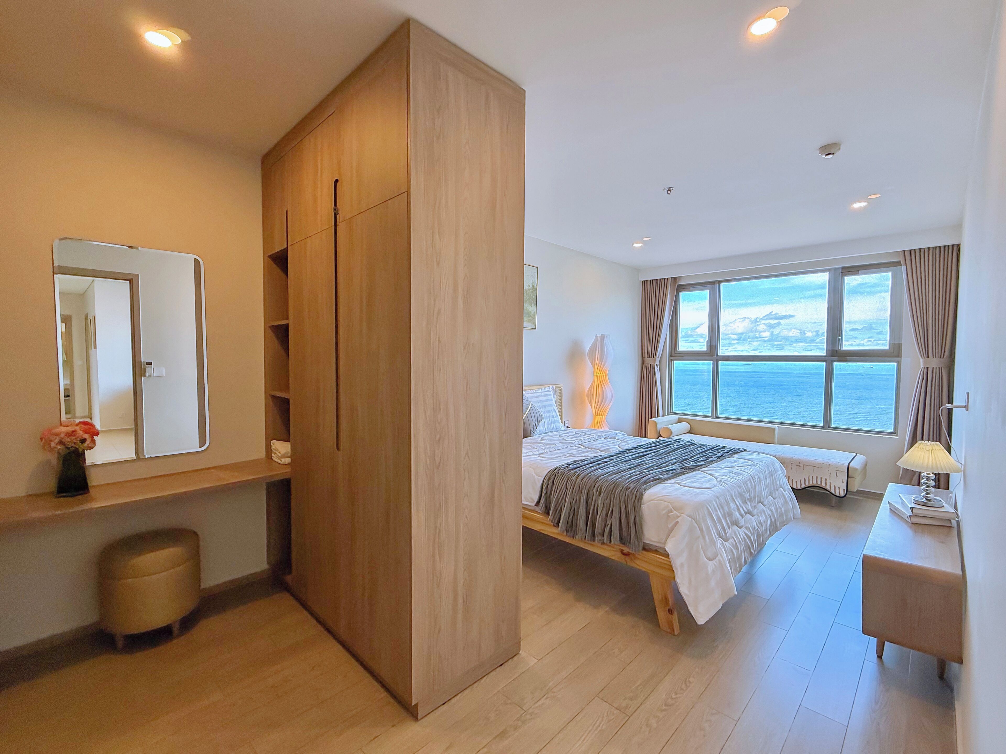 A modern bedroom with a large wardrobe, a bed with a view of the sea, a bedside table with a lamp, and a small stool.