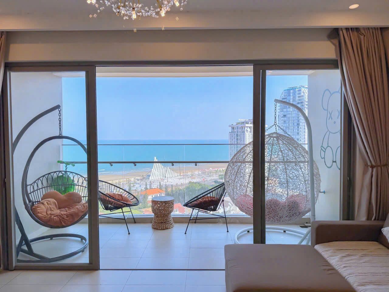 A balcony with a view of the sea and buildings, two hanging chairs, a small round table, and a sofa.