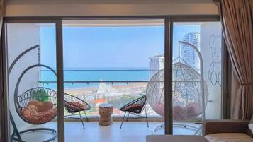 A balcony with a view of the sea and buildings, two hanging chairs, a small round table, and a sofa.