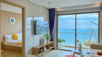 A modern hotel room with a bed, a TV, a wooden console table, a small table with a vase, and a balcony with a view of the sea.
