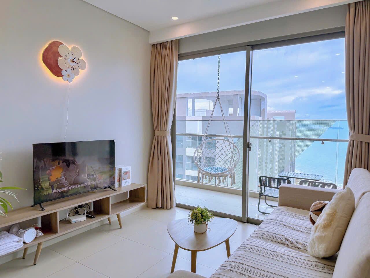 A modern living room with a flat-screen TV, a sofa, a small table with a plant, a hanging chair, and a balcony with a view.