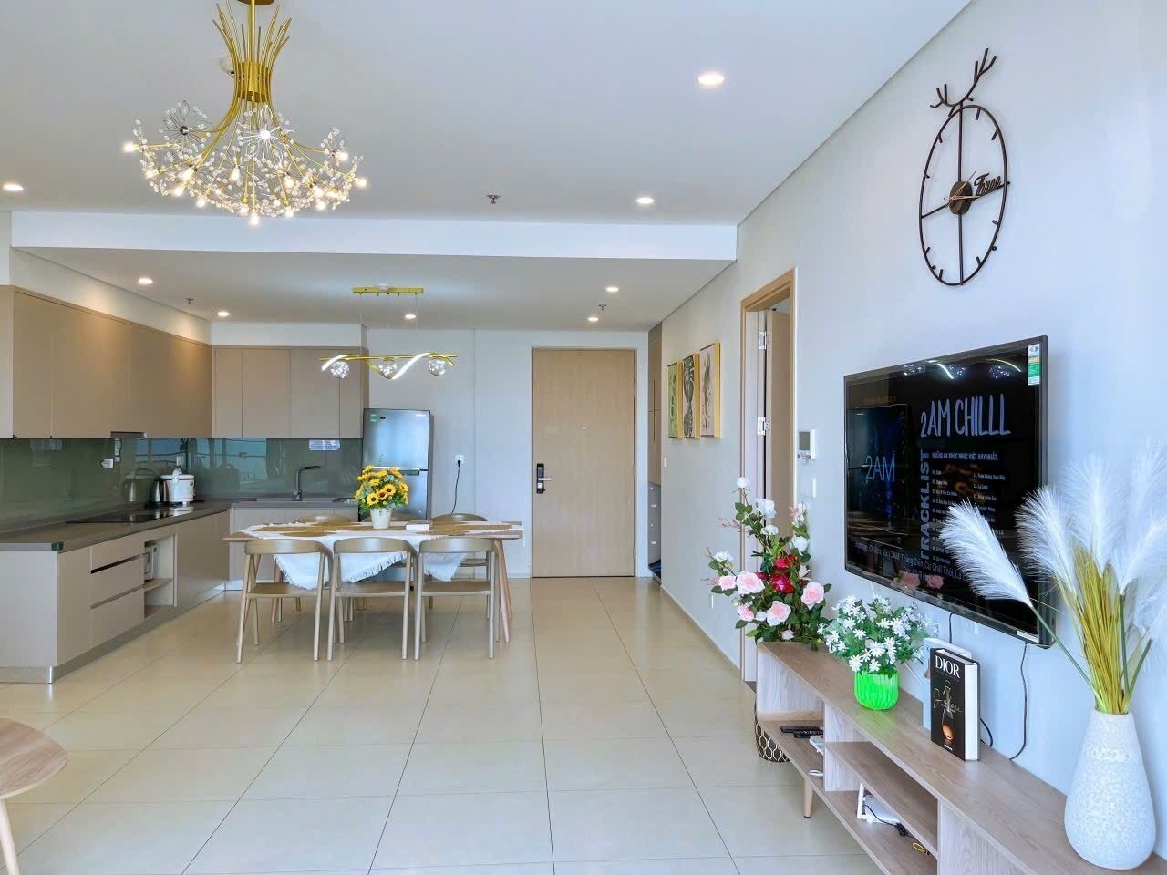 A modern kitchen with a dining area, a TV mounted on the wall, and a decorative clock.