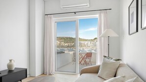 Ein modernes Wohnzimmer mit einer Couch, einem kleinen Tisch und einem Sessel. Ein großes Fenster bietet Blick auf die Stadt und einen Balkon.