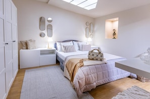 A bedroom with a bed, bedside tables, a nightstand, a desk, a mirror, a wall-mounted shelf, and a skylight.
