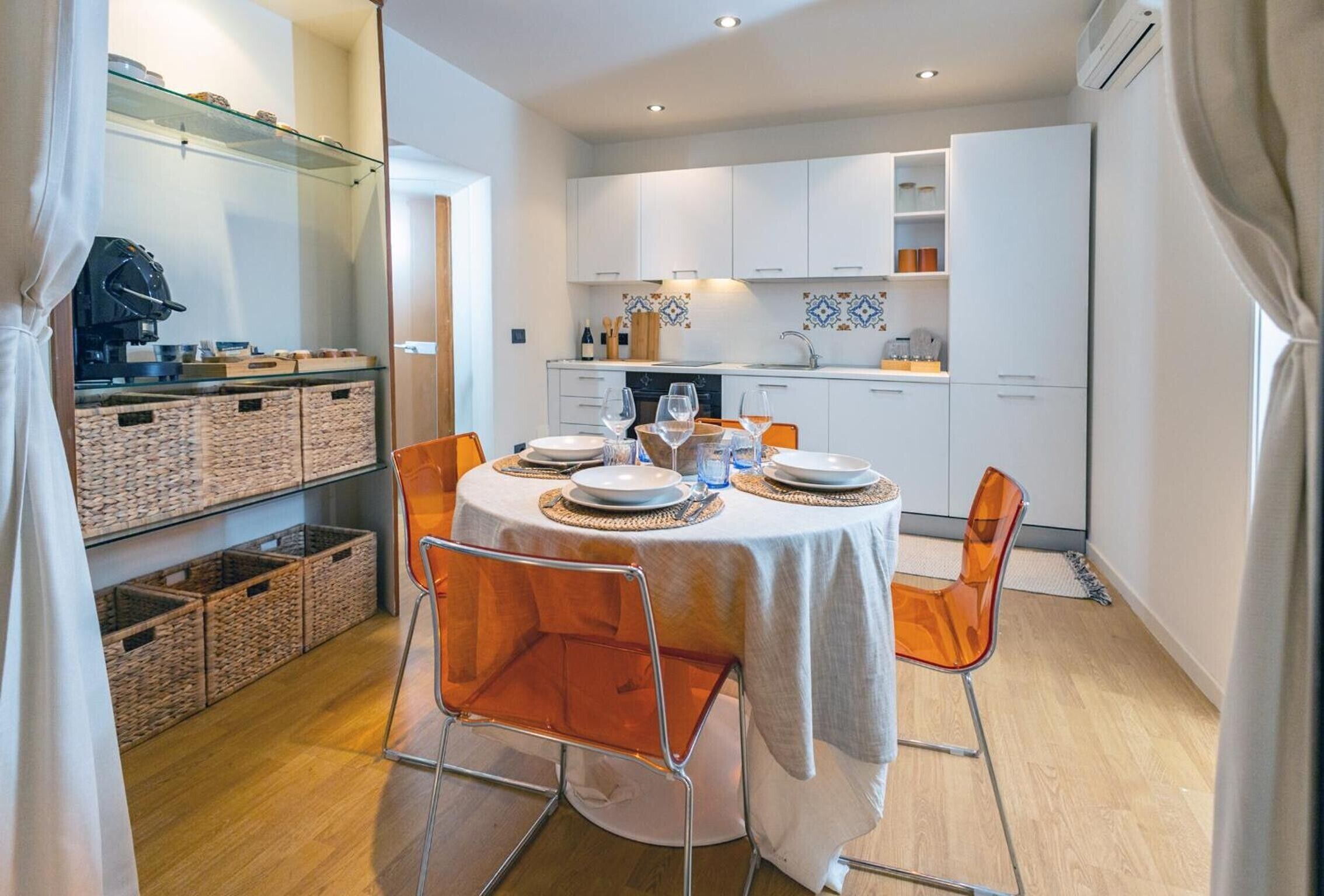 A compact kitchen with white cabinets, a dining table set for four, and wicker baskets on shelves.