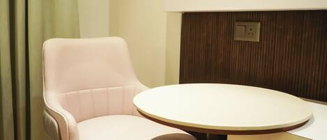 Una habitación con una silla blanca y una mesa redonda, suelo de madera, una cortina verde y un enchufe en la pared.