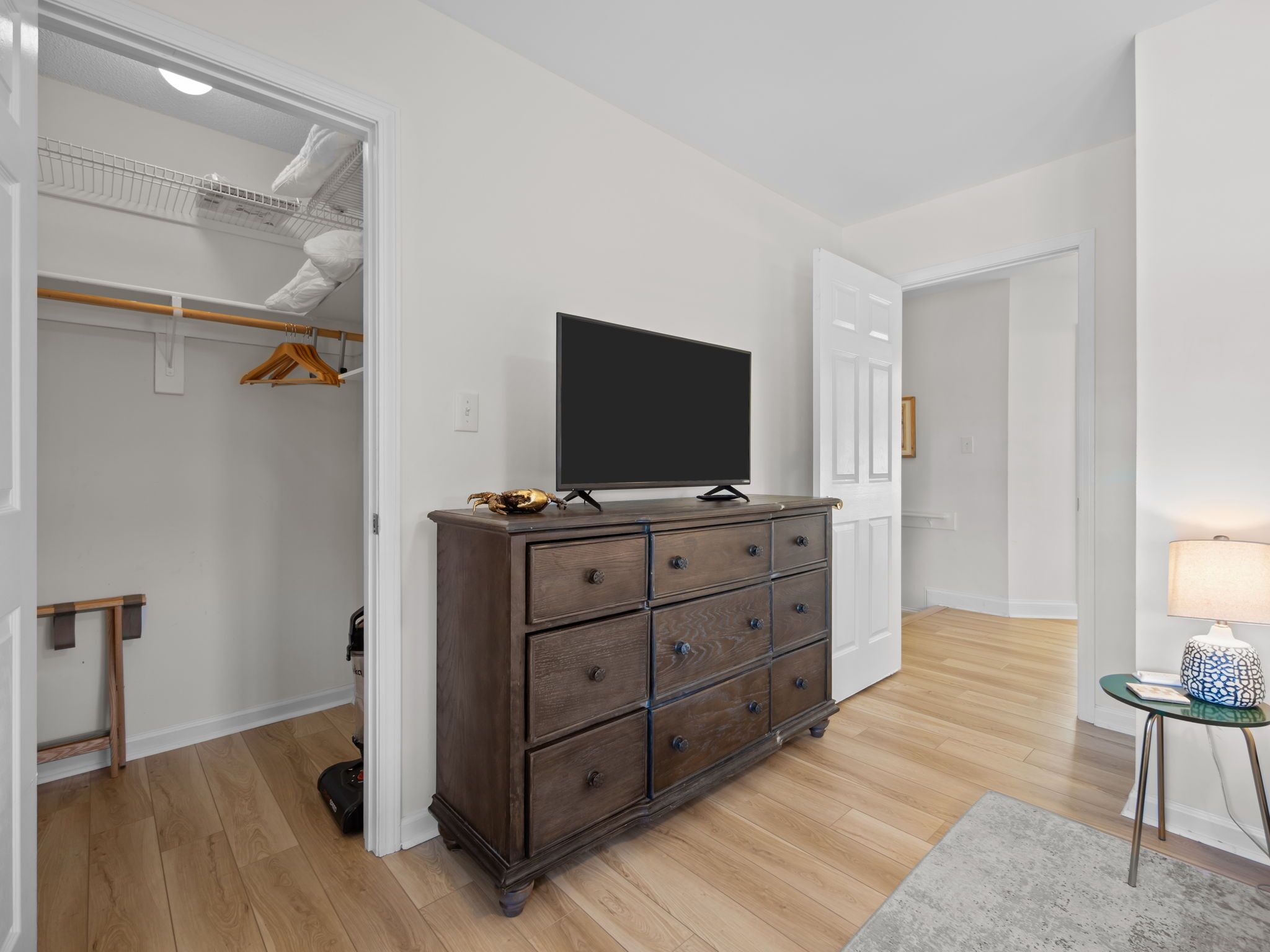 Une pièce comprenant une commode en bois, un téléviseur posé dessus, une armoire avec des vêtements suspendus et une petite table avec une lampe.
