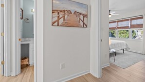 Una habitación con piso de madera, un cuadro de un muelle, una cama con cubrecama blanco y una ventana con persianas.