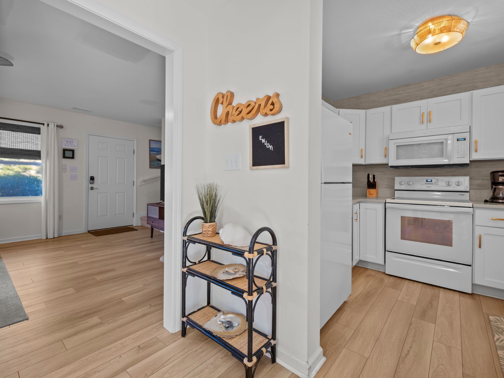 A modern kitchen with white cabinets, a microwave, stove, and a coffee maker. There is a wooden sign on the wall that says "Cheers" and a framed picture with the word "ENJOY".