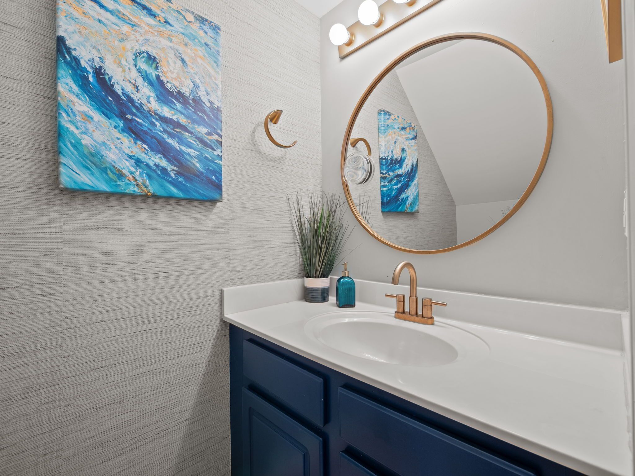 A bathroom with a white sink, blue cabinets, a round mirror with a gold frame, and a blue abstract painting on the wall.