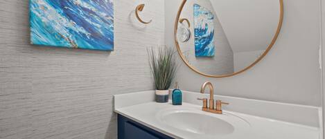 A bathroom with a white sink, blue cabinets, a round mirror with a gold frame, and a blue abstract painting on the wall.