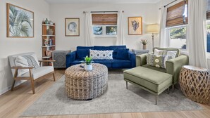 Sala de estar con un sofá azul, un sillón verde y un otomano redondo tejido. Hay un ventilador de techo, una estantería y una ventana con persianas.