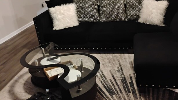 Un salon moderne avec un canapé noir, des coussins à motifs, une table basse en verre et un tapis décoratif.