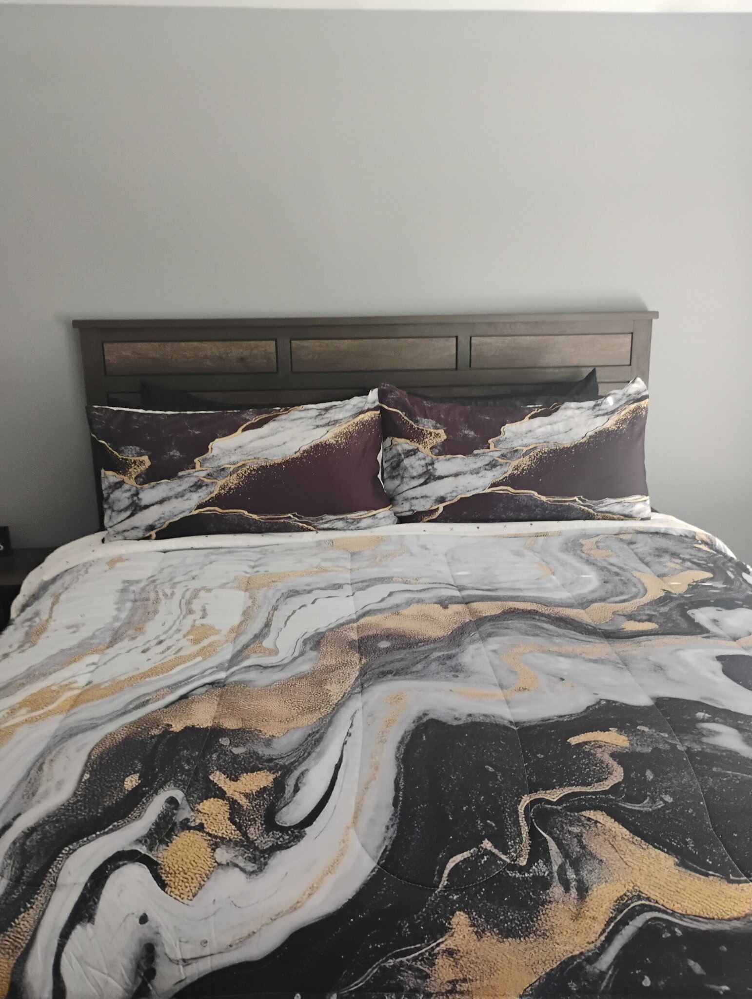 A bed with a marble-patterned bedspread and matching pillows, a wooden headboard, and a plain wall in the background.