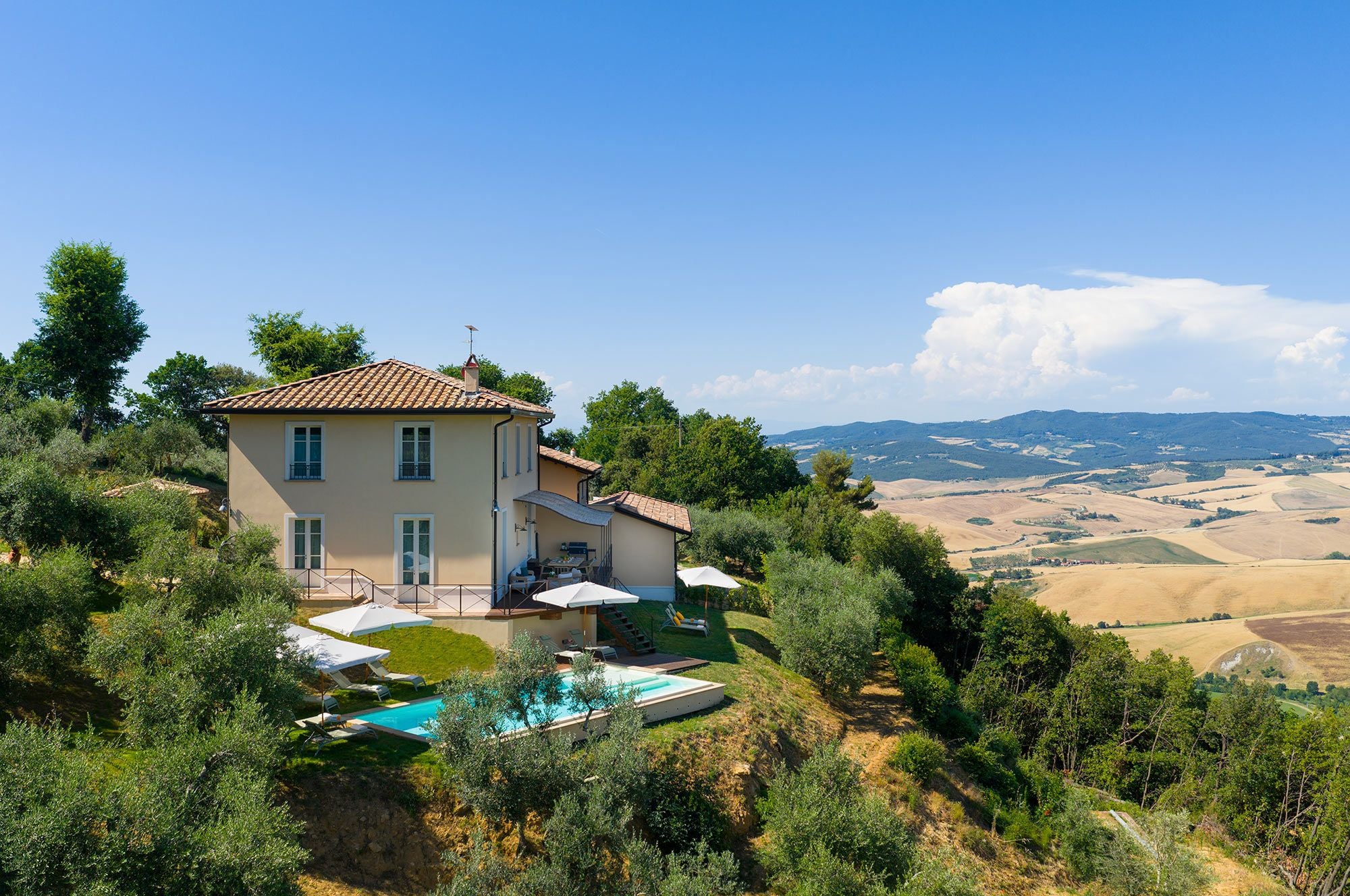 A two-story house with a swimming pool, surrounded by greenery and overlooking a hilly landscape.
