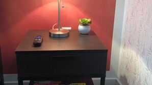 Ein Tisch mit Lampe, Fernbedienung und einem kleinen Topfpflanzen. Darunter ein Magazinregal mit verschiedenen Zeitschriften.