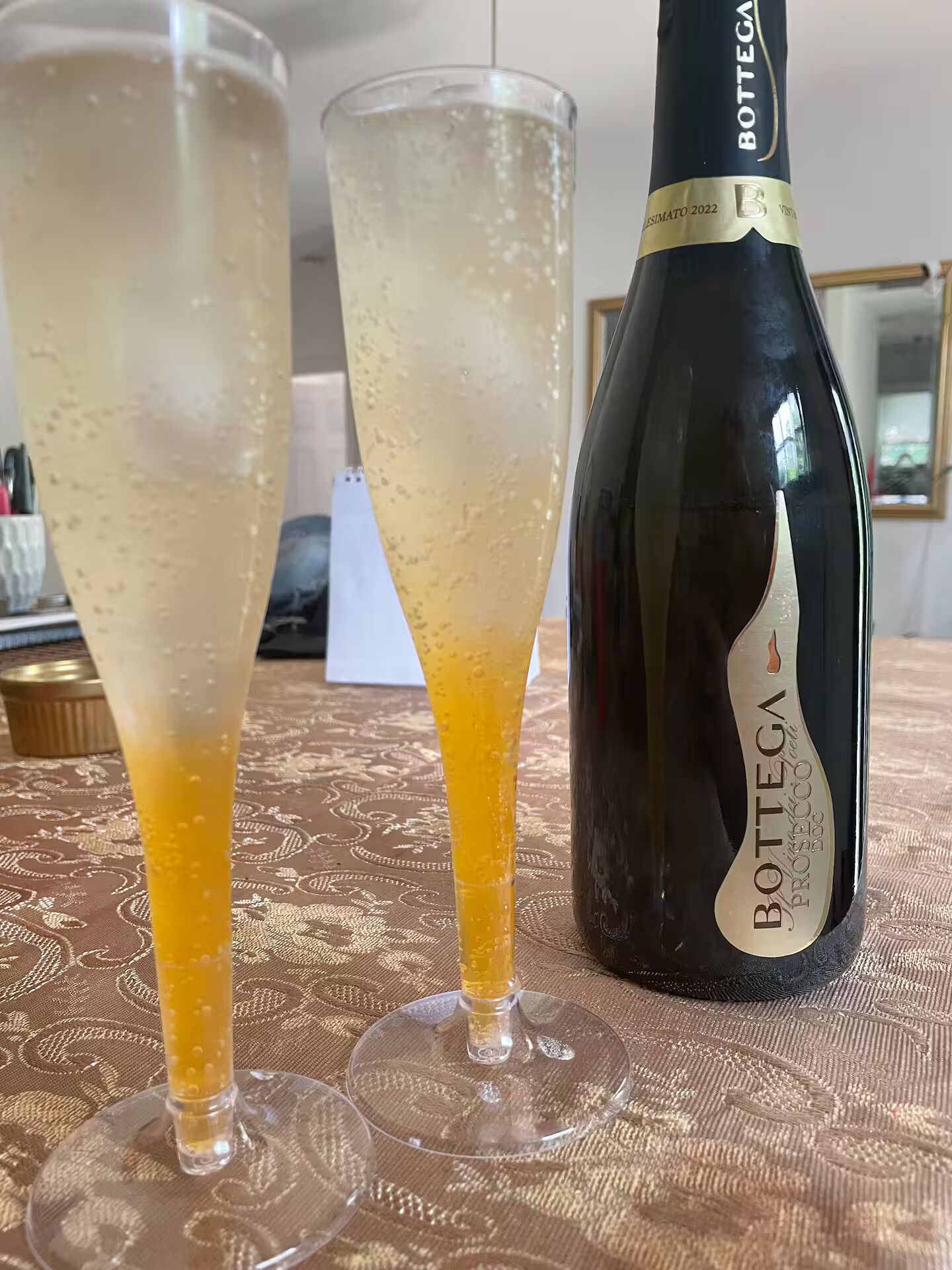 Two glasses of bubbly beverage and a bottle of Bottega Prosecco on a patterned tablecloth.