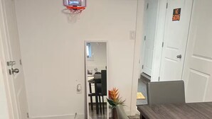 Ein Essbereich mit Holzboden, einem Esstisch und Stühlen. Eine Tür mit einem 'NICHTRAUCHERN'-Schild, ein Basketballkorb und ein Blumentopf sind ebenfalls vorhanden.