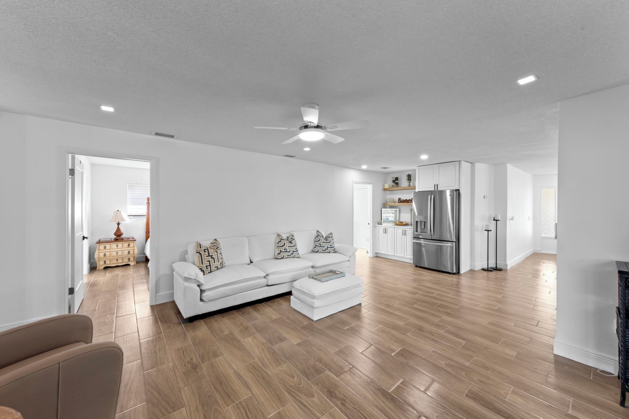 A spacious living room with a white sofa, wooden flooring, and a kitchen area with white cabinets and a stainless steel refrigerator.