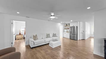 A spacious living room with a white sofa, wooden flooring, and a kitchen area with white cabinets and a stainless steel refrigerator.