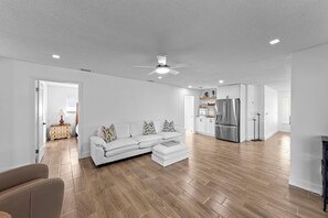 A spacious living room with a white sofa, wooden flooring, and a kitchen area with white cabinets and a stainless steel refrigerator. - Modern riverfront home 4 br & 3 bath with stunning canal water views, private heated pool, garage & private dock access great for fishing, 4 bikes to explore, and minutes to Sanibel, Captiva, and local beaches. (Fort Myers)