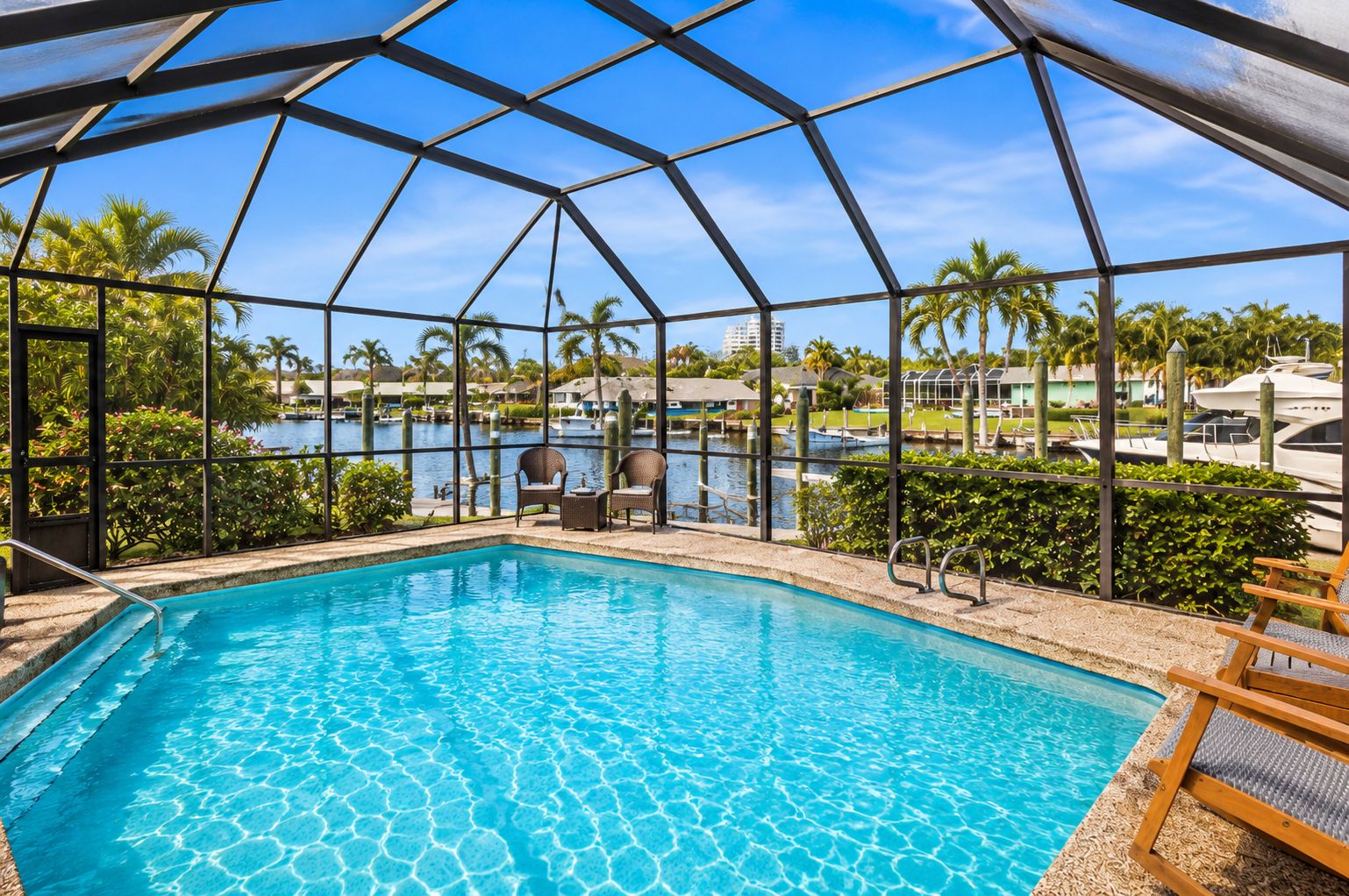 A swimming pool with clear water, surrounded by a metal roof structure, and overlooking a marina with boats and palm trees.