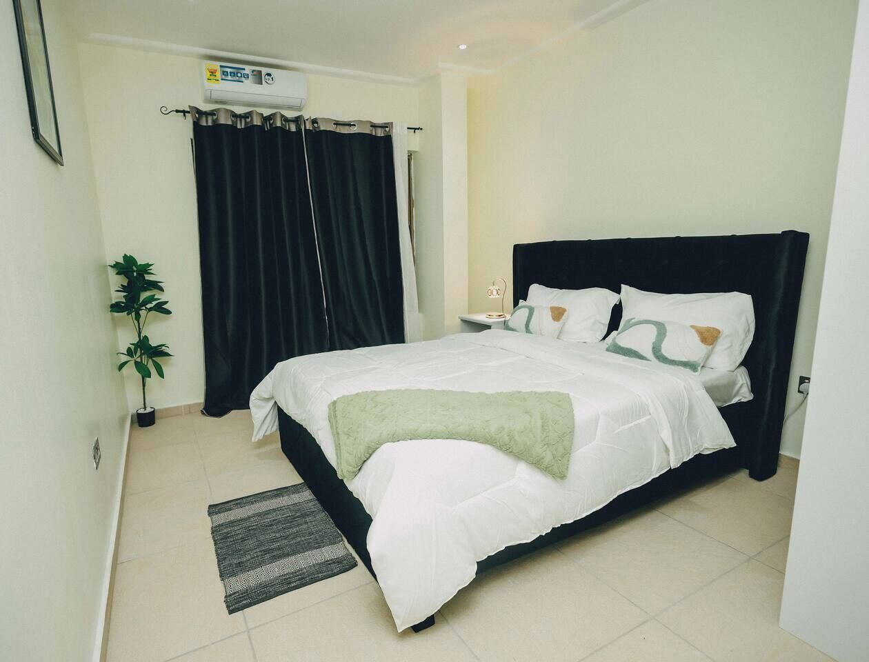 A neatly made bed with white bedding and a green throw, a potted plant, a black curtain, and a wall-mounted air conditioner.