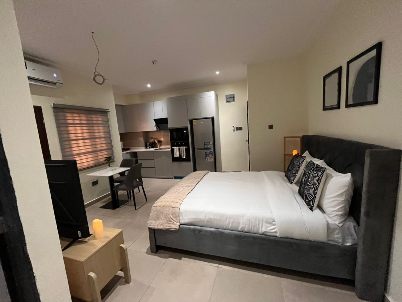 A modern bedroom with a large bed, a desk, and a kitchenette in the background.