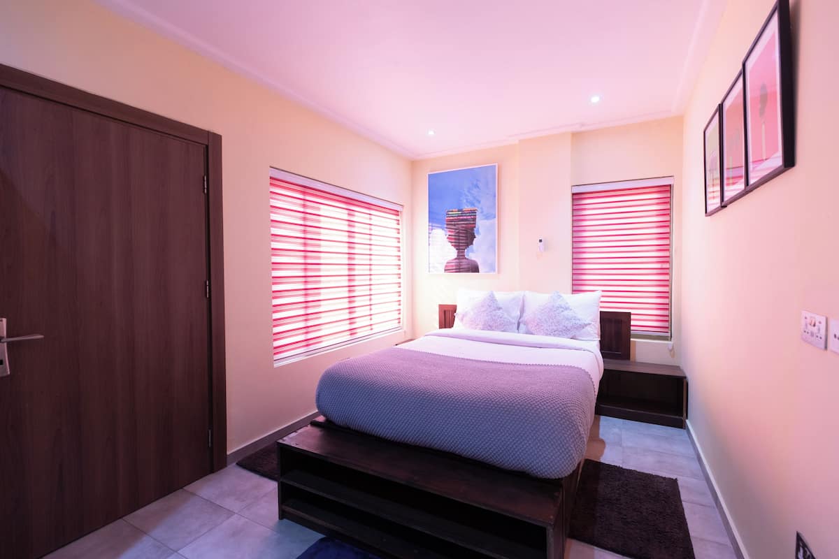 Un dormitorio con cama, dos ventanas con persianas, una puerta y un cuadro en la pared.
