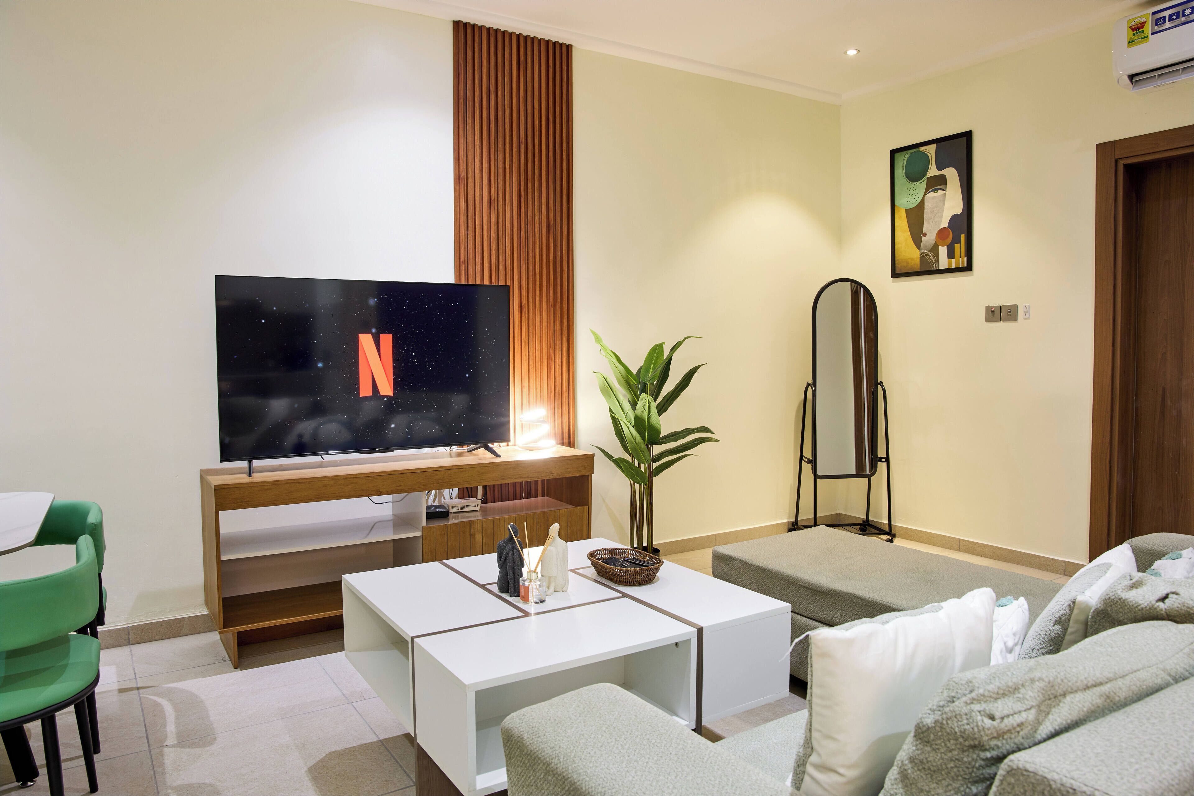A modern living room with a flat-screen TV, a coffee table, a sofa, a green chair, and a potted plant.