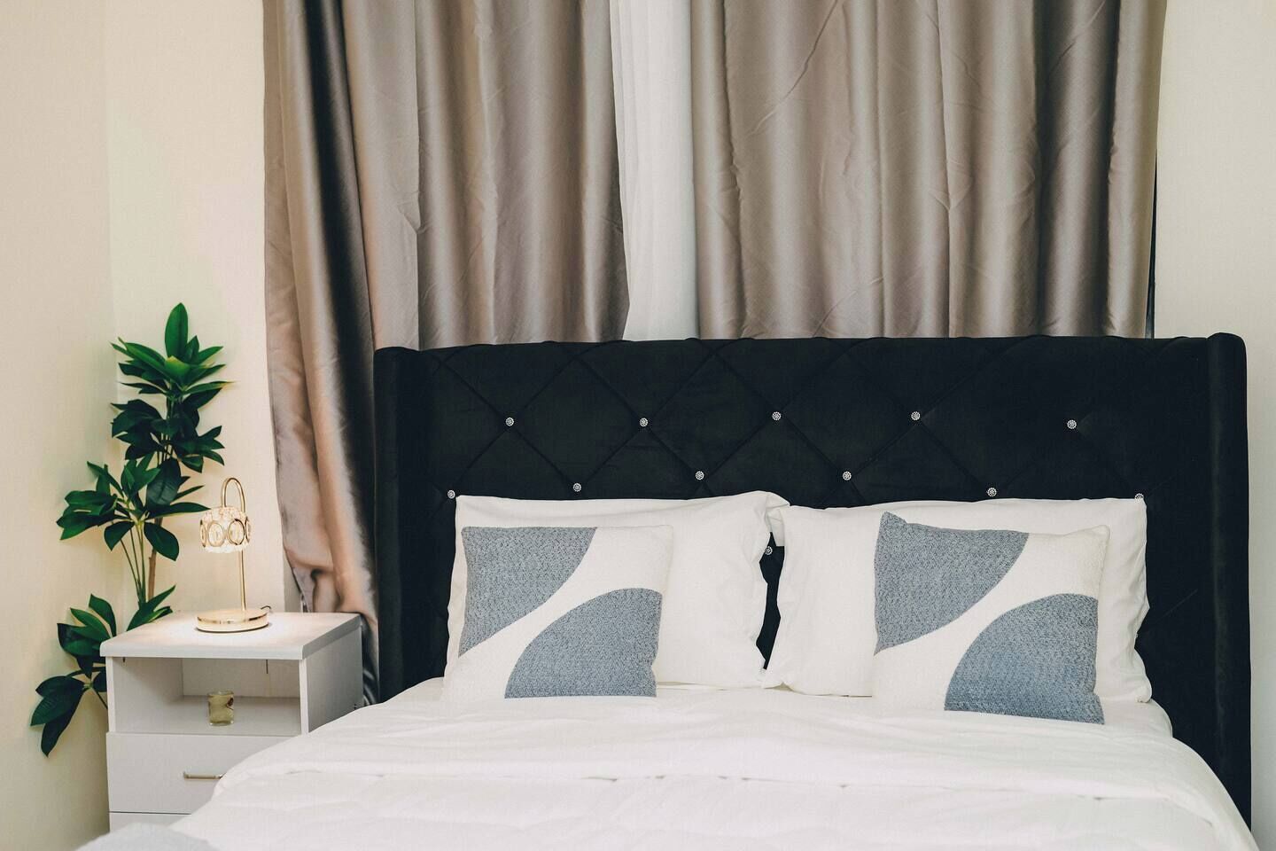 Ein Bett mit schwarzem Kopfteil, zwei Kissen mit geometrischem Muster, ein Nachttisch mit Lampe und eine Pflanze.