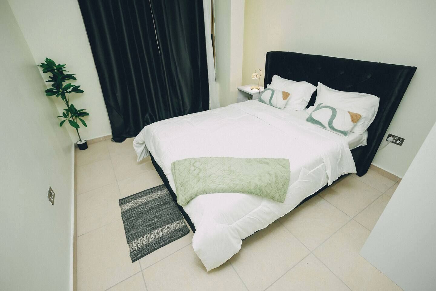 A neatly made bed with a white and green bedspread, a dark headboard, a bedside table with a lamp, and a small potted plant.