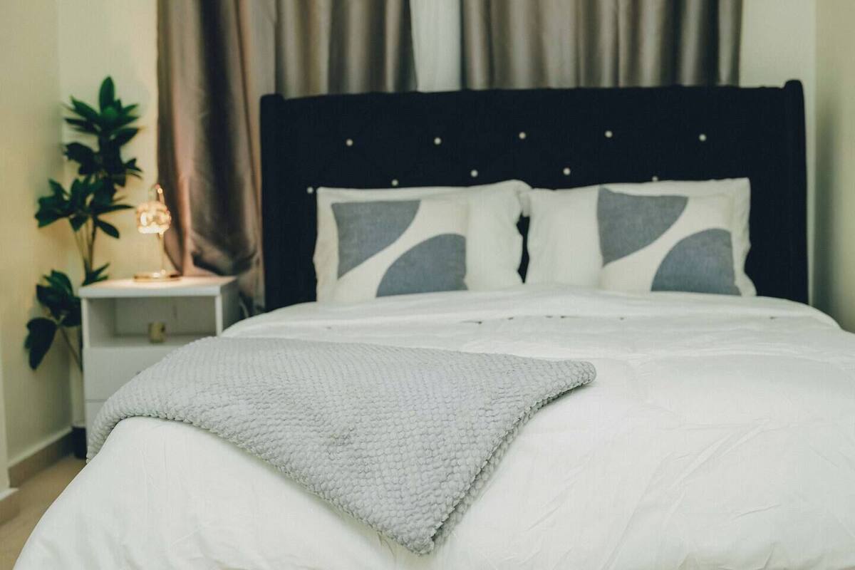 Una cama bien hecha con edredón blanco y manta gris, cabecera oscura y mesita de noche con lámpara.